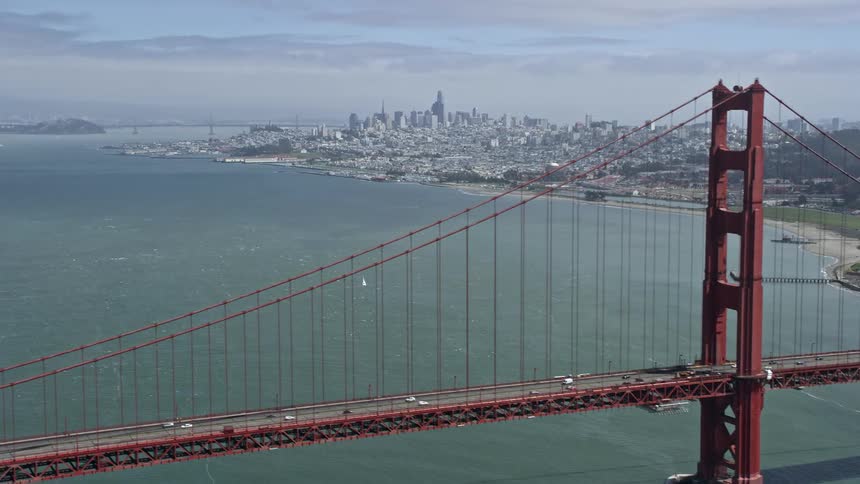 A stunningaerial flyby of the iconic Golden Gate Bridge showcasing the distant cityscape of downtown San Francisco on a partly cloudy day
