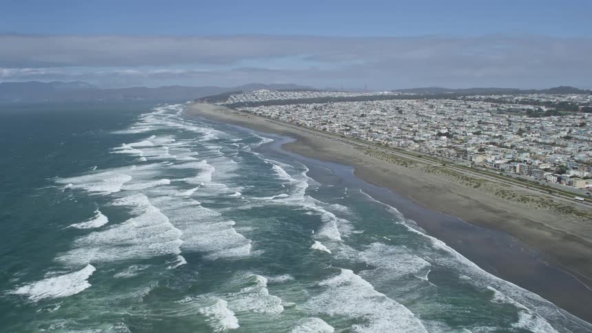 aerial footage captures the beautiful coastline of Ocean Beach and the Outer Sunset District in San Francisco as waves roll towards a partially cloudy shoreline with a wide-angle POV of the residential area and the city skyline in the background showcasi