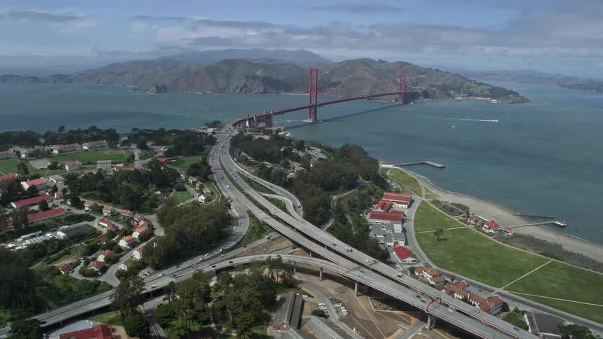 Aerial footage of aresolution clip flying over The Presidio toward the iconic Golden Gate Bridge capturing the historic landmark in beautiful summery daylight with partly cloudy skies and light traffic on the highway