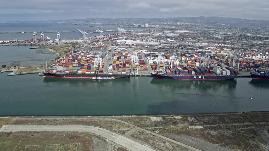 Aerial footage of cargo ships docked at the Port of Oakland in California with cloudy skies and a side view of the vessels