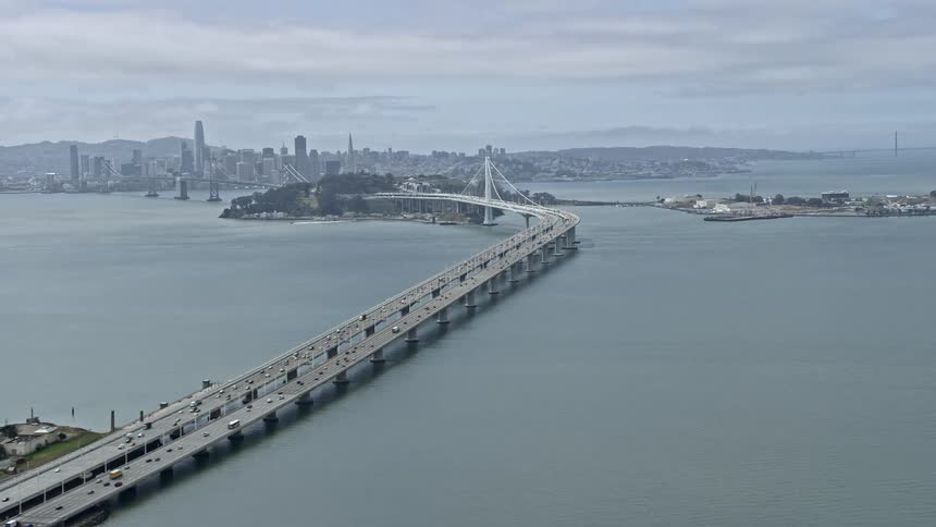  aerial footage of the Bay Bridge in California tilting up to capture the cityscape and downtown area with partly cloudy skies and a wide angle view showcasing the large cable-stayed bridge Interstate 80 and the San Francisco Bay area