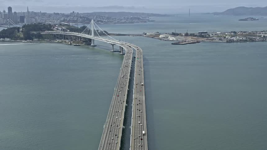  aerial footage capturing the bustling traffic on the Bay Bridge as it stretches across San Francisco Bay with a magnificent cityscape in the background on a partly cloudy day showcasing the iconic suspension bridge in the downtown area of San Francisco C