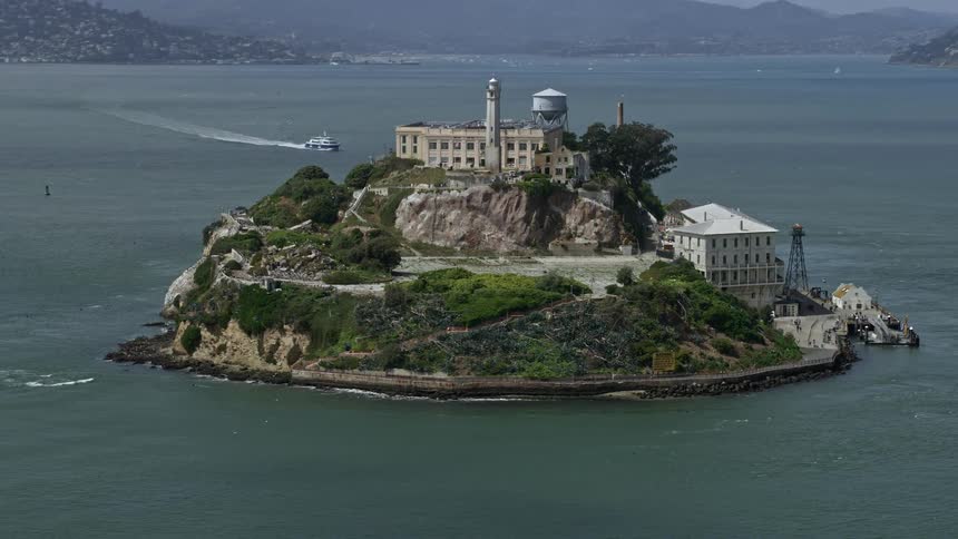  aerial footage captures a view of Alcatraz Island a historic government building and iconic correctional facility as the camera flies away over the San Francisco Bay on a partly cloudy day in Northern California