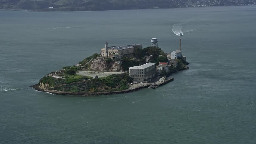 Aerial footage flying towards Alcatraz Island a historic government building and iconic structure located in San Francisco Bay California with partly cloudy skies and beautiful views of the bay area and west coast captured using a Shotover F1 camera syste