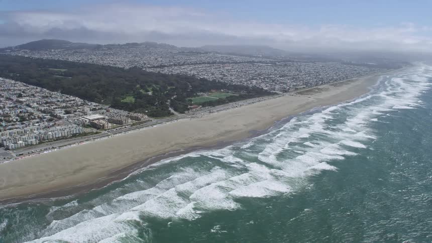 aerial footage capturing an reverse view of Golden Gate Park Ocean Beach and the Outer Sunset district in San Francisco California showcasing the city's iconic shoreline city park and residences during a partly cloudy day