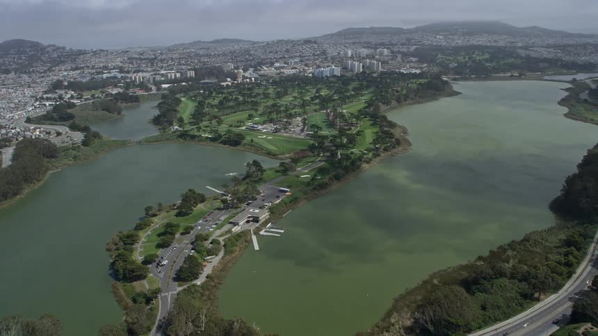  aerial footage of a golf course and Lake Merced in Lakeshore San Francisco California showcasing a beautiful view from above as the camera approaches the scenic area with partly cloudy skies and the city in the distant background