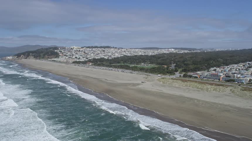 anaerial footage showcasing the stunning Ocean Beach and Golden Gate Park in San Francisco California panning from the ocean with partly cloudy skies and waves crashing onto the empty beach capturing the iconic structures of the city while approaching the