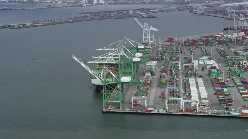  aerial footage capturing a cargo ship passing under cranes at the Port of Oakland California with cloudy skies and the bustling activity of the shipping industry