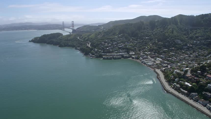 Thisaerial footage showcases the scenic Sausalito neighborhoods as the camera flies towards the iconic Golden Gate Bridge in the distance with the California shoreline and partly cloudy skies as the backdrop