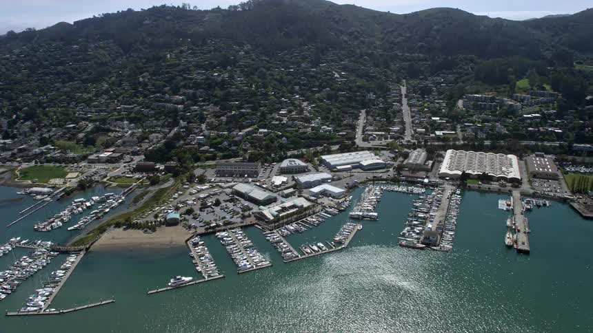 Aerial footage of Sausalito marinas in California during the summer showcasing the seaside harbor with boats docks residential areas and a town on a slope above the marina under partly cloudy skies
