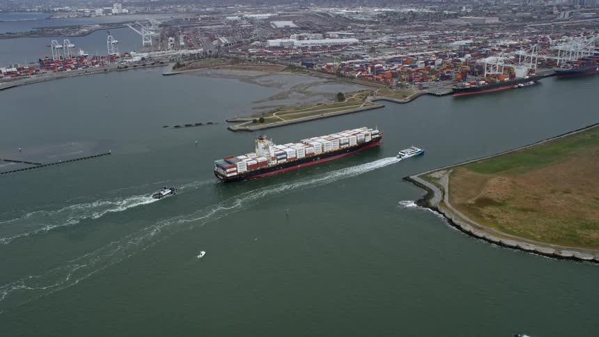  aerial footage captures a cargo ship and escort sailing near the Port of Oakland California amidst cloudy skies and bustling harbor activity