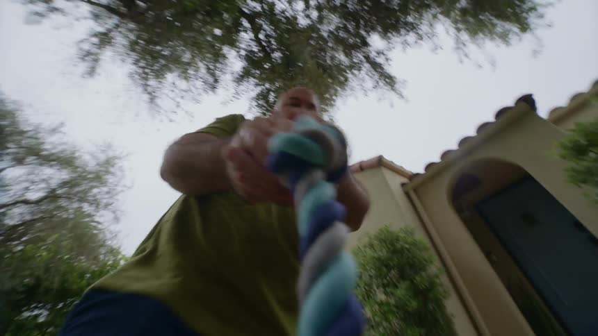 Man outdoors playing with a rope toy as if holding it from the mouth of a dog outside the frame during day time