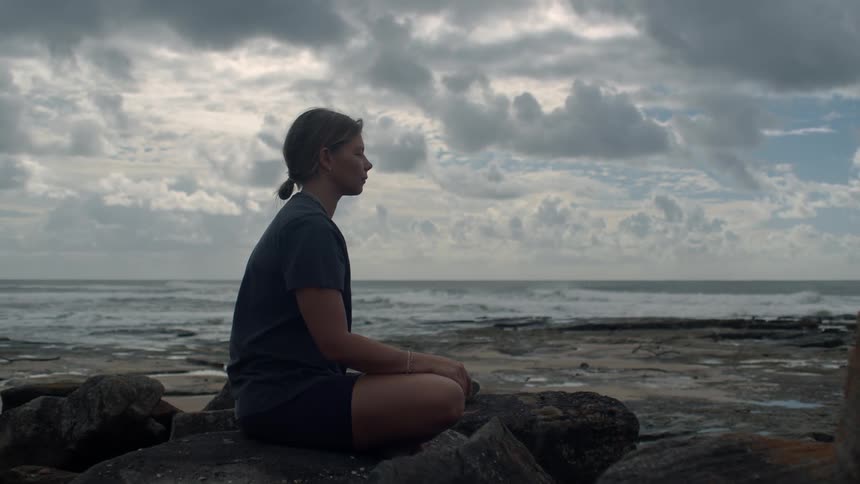 A woman mediates on the rocky shore of the ocean during the day