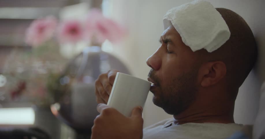 A man in pain with a towel on his forehead drinks from a cup while sitting in bed