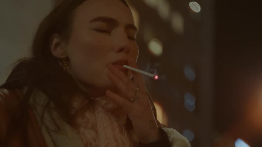 A woman standing outdoors at night concentrates on lighting her cigarette