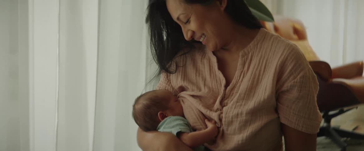 A woman breastfeeds a child while rocking the child in her arms indoors during the day