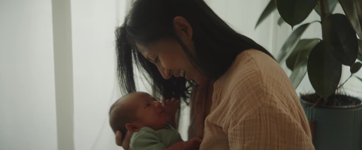 Mother holding her newborn baby in her arms and to her chest standing in front of a shaded window as the sunlight shines through