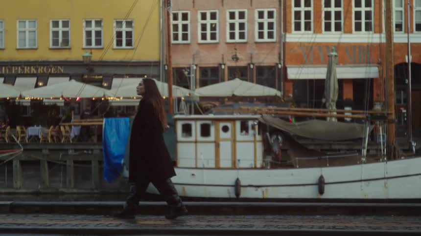 A woman walks on a sidewalk and looks around next to a boat on a canal and buildings in a european town