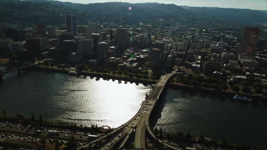 Fly over Morrison Bridge and Willamette River towards Downtown Portland capturing the high-rise buildings and city square under the bright sunshine of a summer day