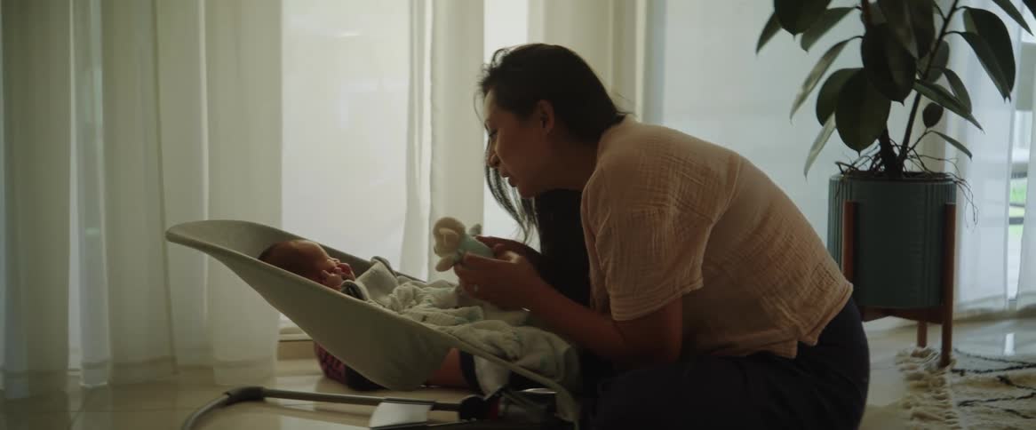 A mother sitting on the floor in front of her newborn baby in a seat in front of her while she holds a stuffed toy in front of the babies face