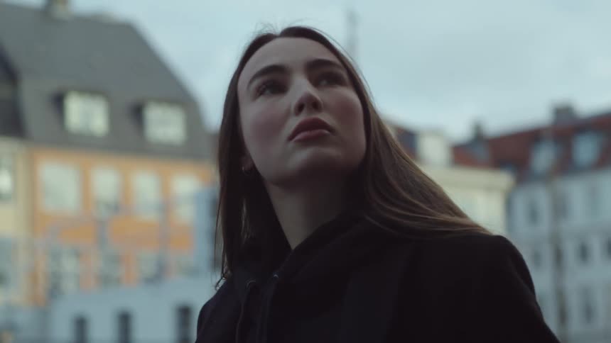 Close up of a woman wearing a black coat walking outside near city buildings as she looks up and around her