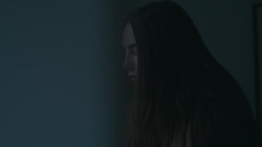 A woman with long brown hair sits in a dark room looking contemplative