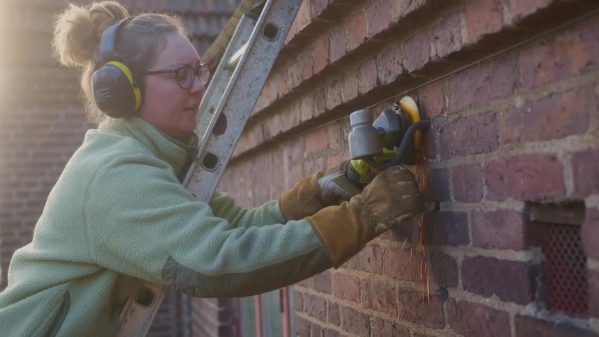 A woman on a ladder uses a circular saw to remove an outdoor sconce lamp