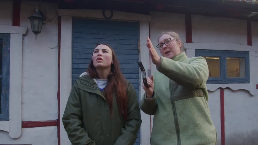 A woman explains something to another woman outside a house while holding a metal object and gesturing