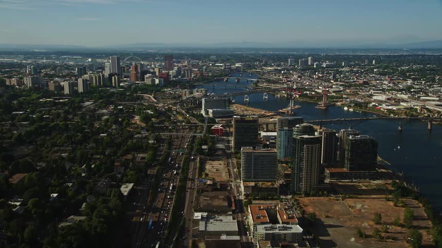  aerial footage showcasing the bustling highway passing by towering highrise buildings as it runs alongside the picturesque river leading towards the vibrant downtown area of Portland Oregon