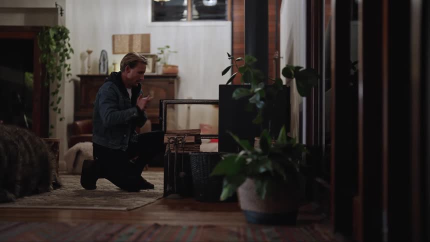 A man crouches down putting wood on a fireplace to keep warm and rubs his hands together