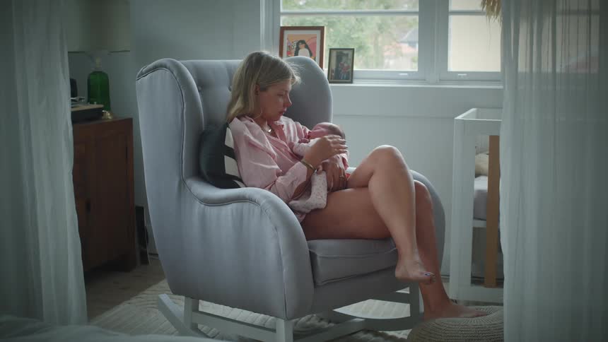 Woman with nervous expression sitting on an armchair with a sleeping baby on her arms during day time