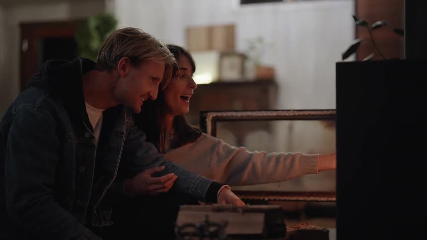 Man and woman sit close to fireplace as she feeds fuel into flames