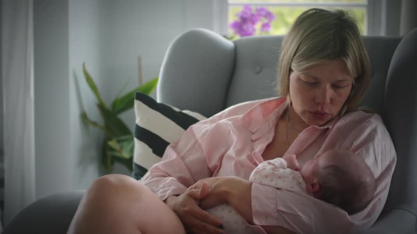 A white woman sits on a chair gently rocking back and forth holding a baby in her arms and wearing a wired earbud
