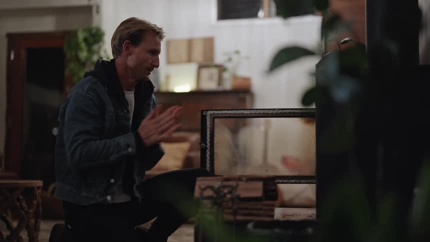 A man in a jacket rubs his hands together while putting wood into a furnace