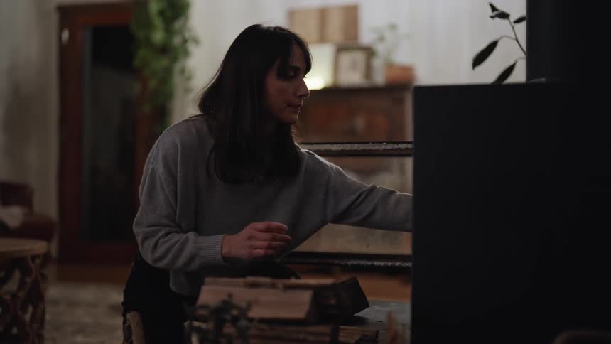 Woman standing up indoors adding wood to a fireplace during day time