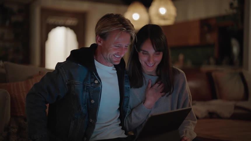 A man and a woman sitting on a couch look at a tablet and laugh
