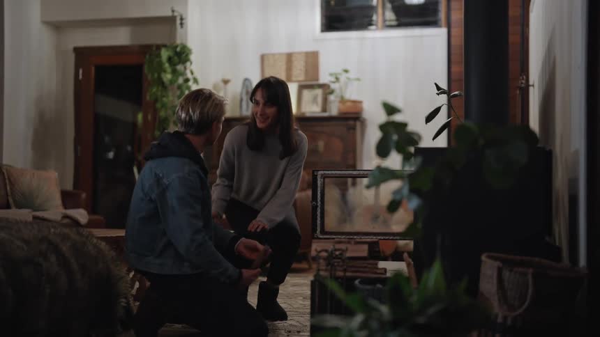 A woman walks into a living room carrying a basket of wood where a man is waiting for her kneeling by the fireplace greeting her with a kiss