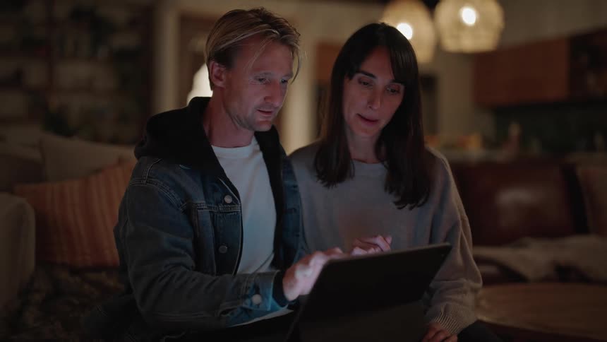 A man and a woman sitting on a couch look intently to a laptop monitor as the man writes on the keyboard