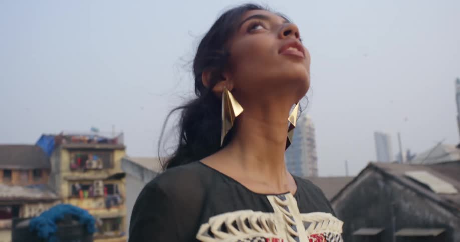 A young woman wearing gold earrings with her long dark hair pulled back looks around as she walks through a neighborhood and birds fly overhead