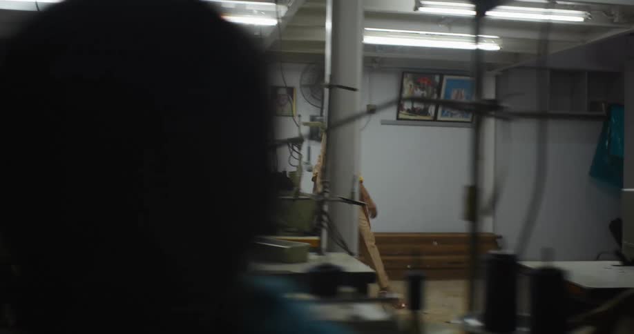 A woman walks by a production line where two men use sewing machines before exiting