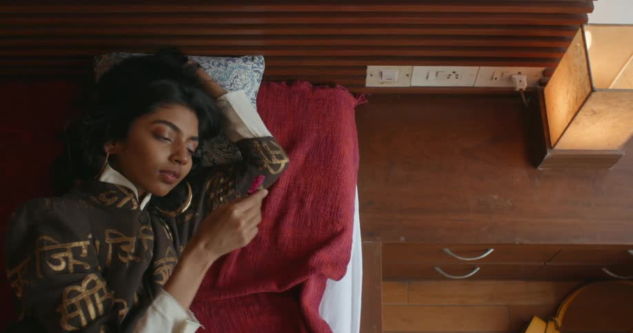 Woman lying on bed picks up flower head lying on dresser and examines it