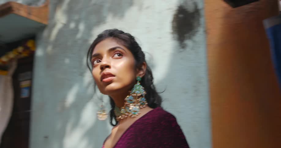 A woman with elaborate earrings and necklace and bohemian clothes emerges from a building and runs away from the camera down an alley