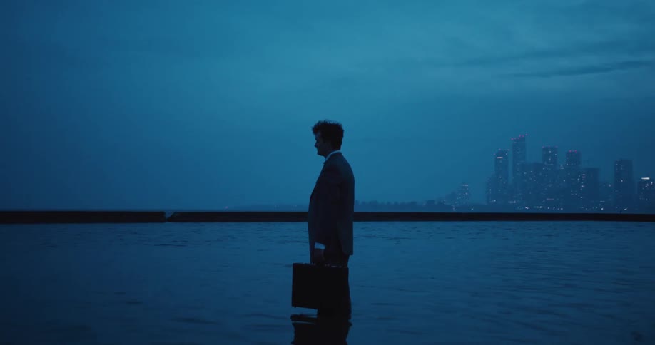 Man in a suit holding briefcase stands up to knees in water with skyline in the distance at blue hour