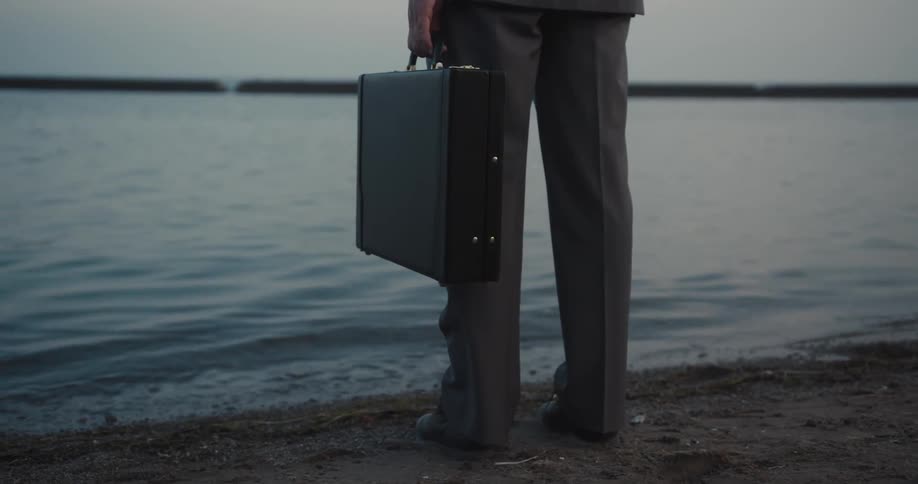 A pensive businessman holds a briefcase at the lakeside in the evening
