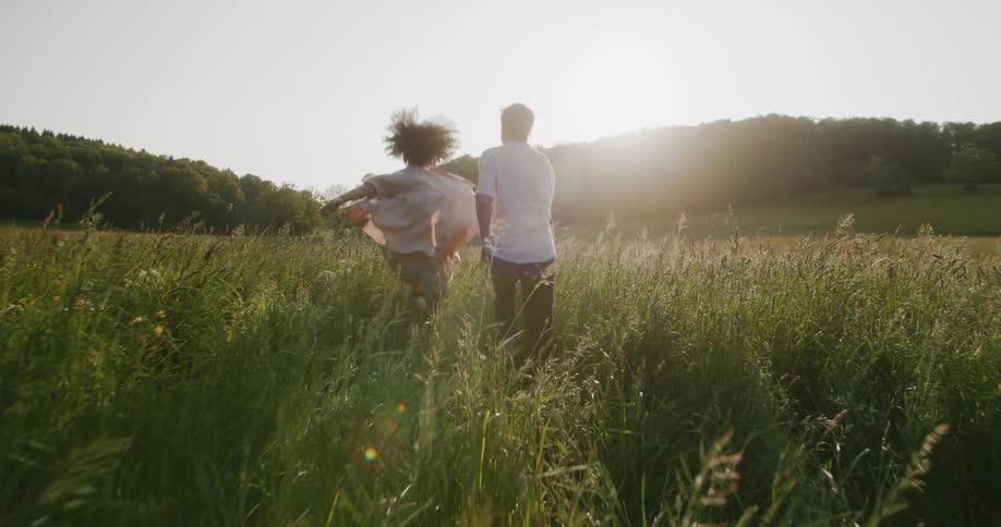 A woman and a man running through tall grass in a field as the sun is falling behind tree covered hills