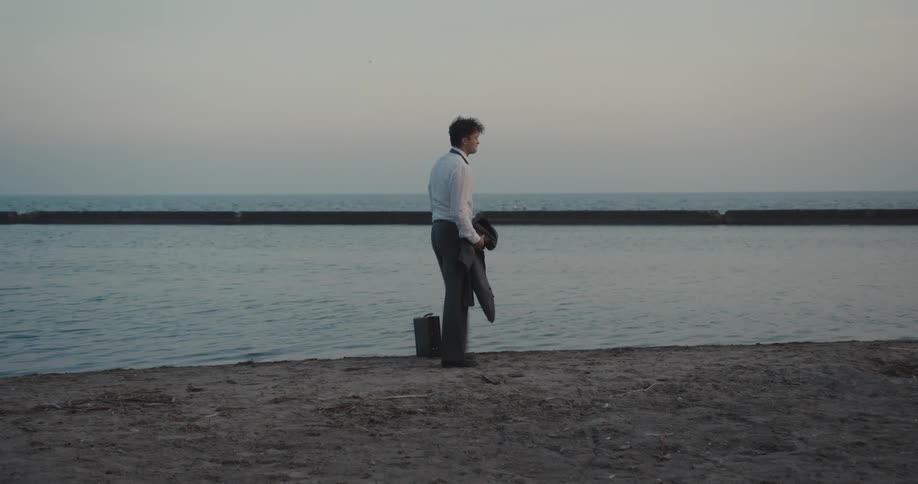 A man in a suit on the edge of the sea walking then kicks the sand and puts one hand on his head