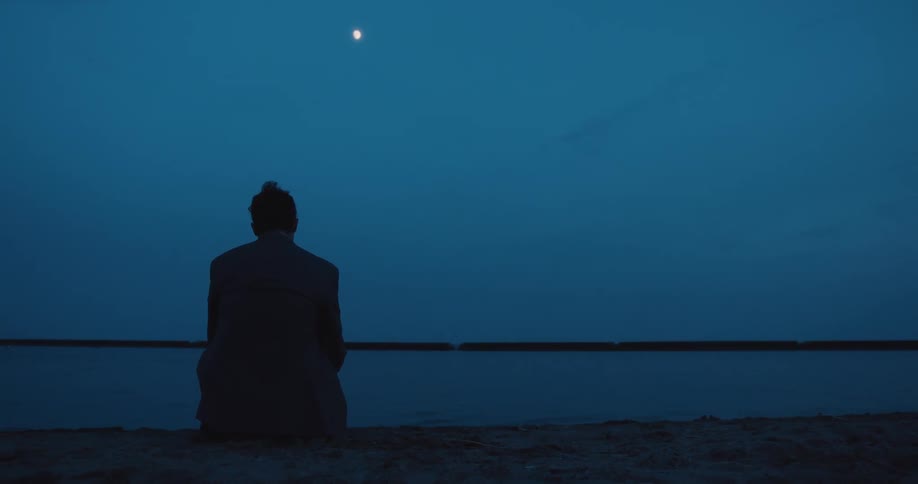 A distraught businessman sits on the edge of the lake watching the moon at night