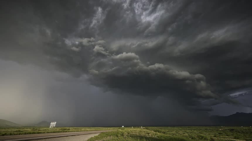 A huge storm cloud fills the sky with gray next to a highway with several cars passing by