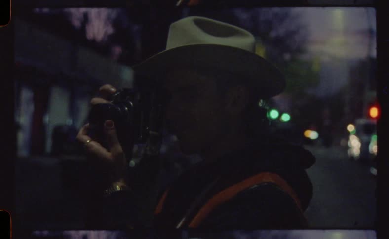 A man in a hat in the middle of the street taking pictures with a digital camera
