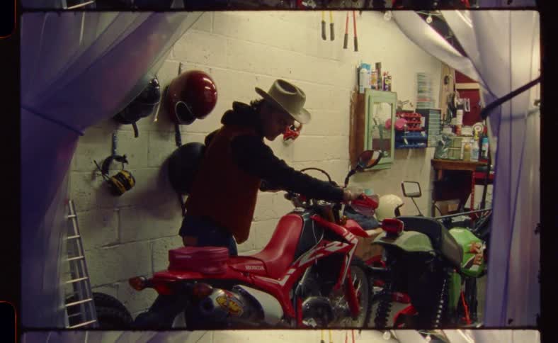 A man walking a dirt bike backward out of a small garage area with plastic folds held back for the door